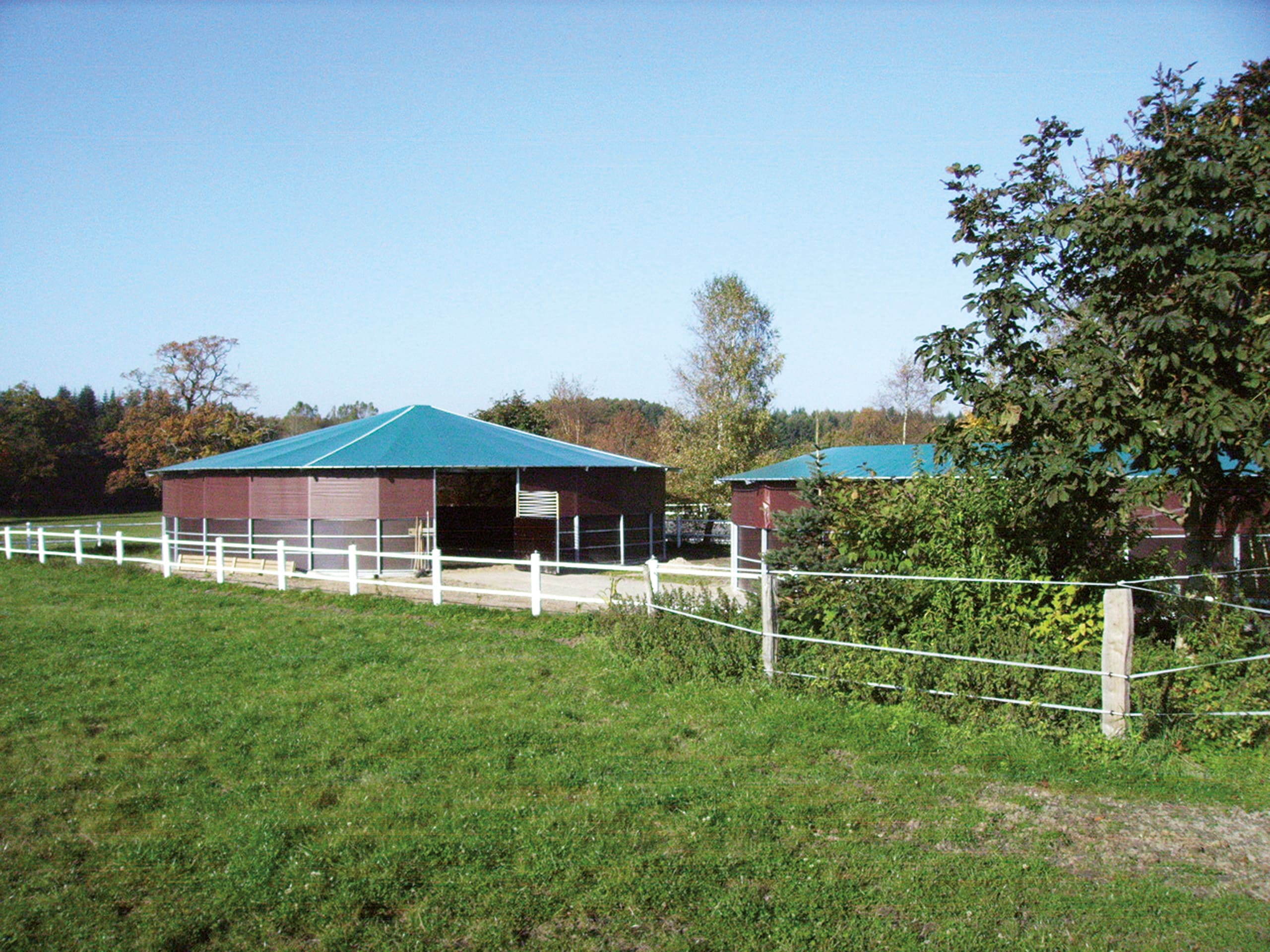 Sicht über Hofgelände und Koppel, in der Ferne eine Longierhalle mit braunem Windschutznetz