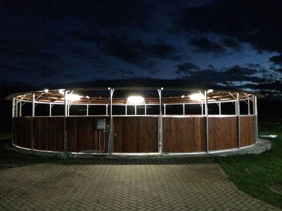 Beleuchtete Longierhalle mit Holzverkleidung bei Nacht von außen